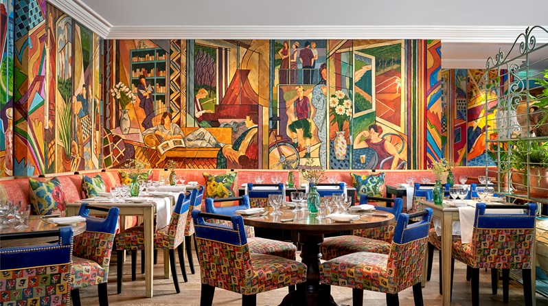 Oscar Restaurant tables with white table cloths, glasses and fresh flowers on each. A colourful mural covers the restaurant walls.