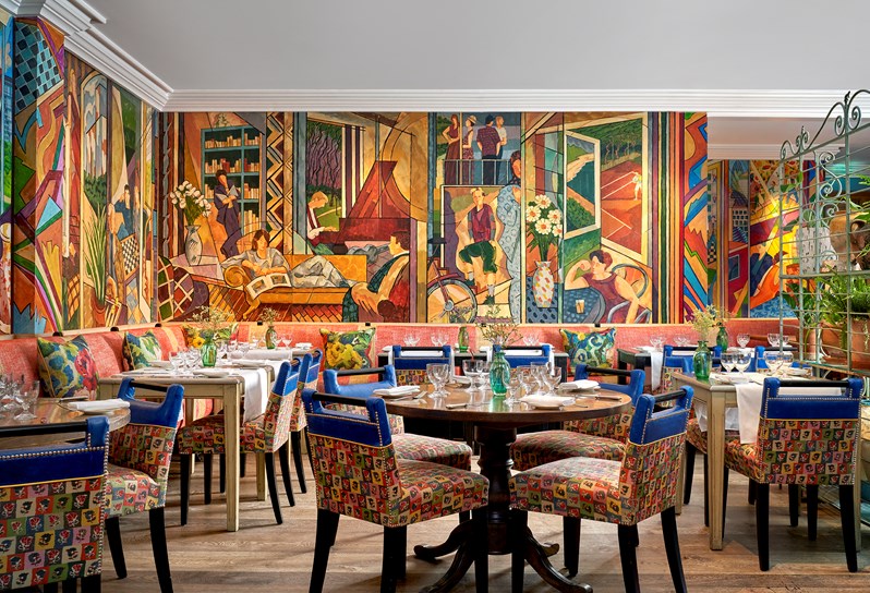 Oscar Restaurant tables with white table cloths, glasses and fresh flowers on each. A colourful mural covers the restaurant walls.