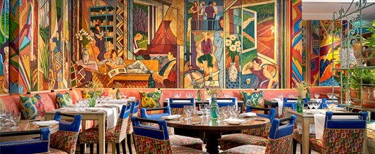 Oscar Restaurant tables with white table cloths, glasses and fresh flowers on each. A colourful mural covers the restaurant walls.