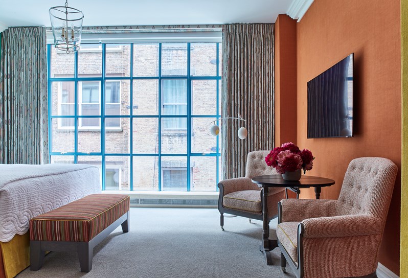Two striped armchairs, a coffee table with pink and red flowers and a TV in front of an orange wall. Floor to ceiling windows and the end of a bed with a striped ottoman.
