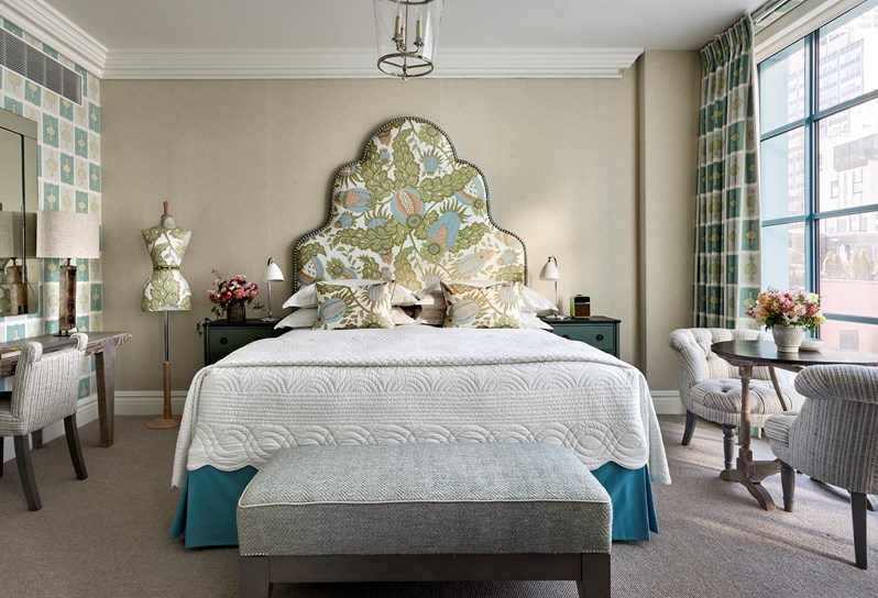 A bed with a headboard upholstered in a pale blue, green and pink floral pattern on a white background, a matching mannequin next to it and a grey ottoman at the end. Two striped blue chairs and a coffee table in front of the window which have curtains in Ozone fabric in a blue colour scheme, which matches the wall opposite, with a desk and white chair in front of it.