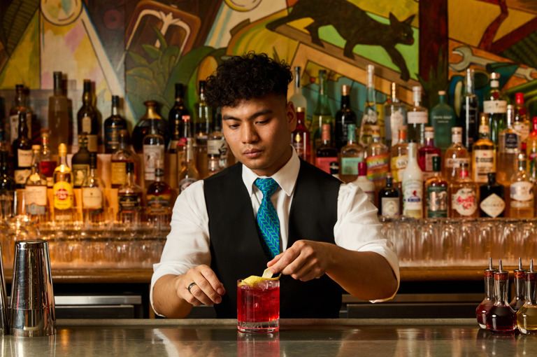 An image of a male bartender making the Ruby Reverie cocktail in the Refuel Bar, at Soho Hotel. The image shows the bartender placing the lemon rind glass on the glass.