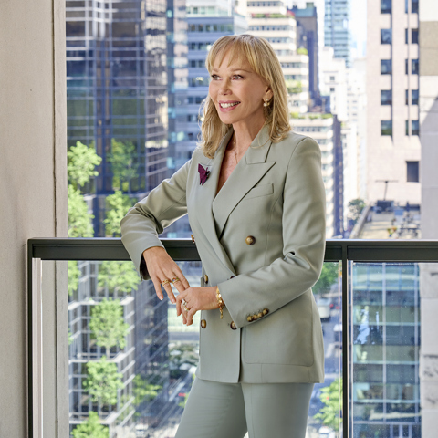 An image of Kit Kemp at Warren Street Hotel. She is smiling and looking off camera in a suit, standing in front of a window that showing Tribeca in the Background