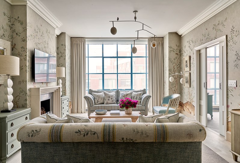 A neutral coloured floral patterned sofa, cushions and wallpaper opposite a blue striped sofa, a wooden coffee table in the middle in front of fireplace with a TV above.
