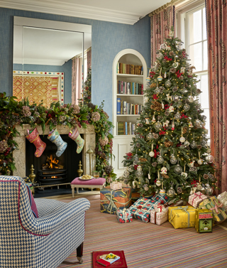 This image shows a beautifully decorated Christmas setting in the townhouse at Haymarket Hotel, featuring a Christmas tree adorned with twinkling lights, red berries, pinecones and ornaments, surrounded by colourful wrapped presents. A roaring fireplace is dressed with festive foliage and stockings, completing the cosy seasonal scene.