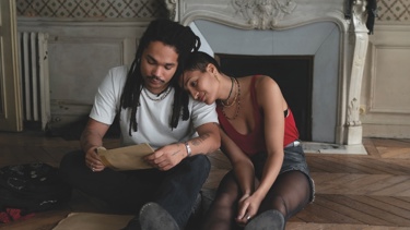 A still from the film "Father Mother Sister Brother": A young woman leans on a man's shoulder as they both look at a letter, sat down in front of a fireplace