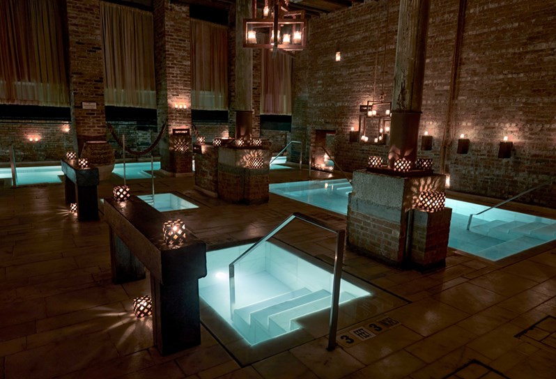 A subterranean space with exposed brick walls and a number of glowing plunge pools bathed in soft candle light