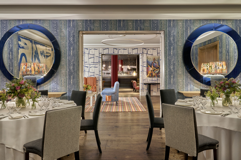 An image of multiple round dining table set up in the Indigo Room, there are white table cloths, floral bouquets in the centre and candles. each place around the table has a placemat, plate, cutlery, glassware and napkins ready for dining. private events room at The Soho Hotel.