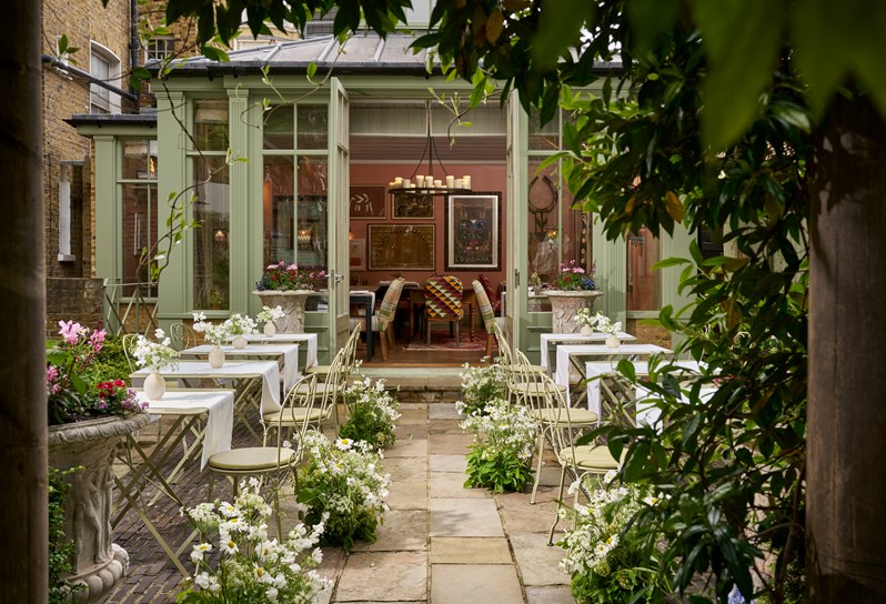 Shot from under a stone archway. The camera looks down a path in the middle of a patio, there are tables of two set up either side. At the end of the patio, the doors to The Orangery are open.