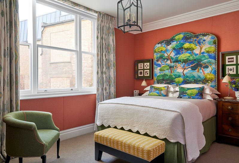 A view of the bed with a color tree headboard and white linen. The walls are red colour and there is a green armchair to the right of the window and a yellow ottoman at the bottom of the bed.