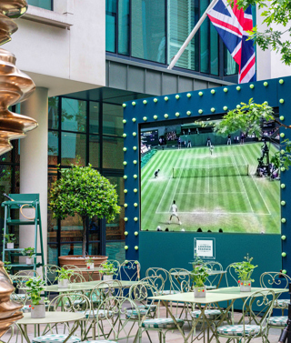 An image of the big screen set up on the Ham Yard Hotel's courtyard displaying Wimbledon, with green tables and chairs in front of it