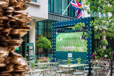 An image of the big screen set up on the Ham Yard Hotel's courtyard displaying Wimbledon, with green tables and chairs in front of it