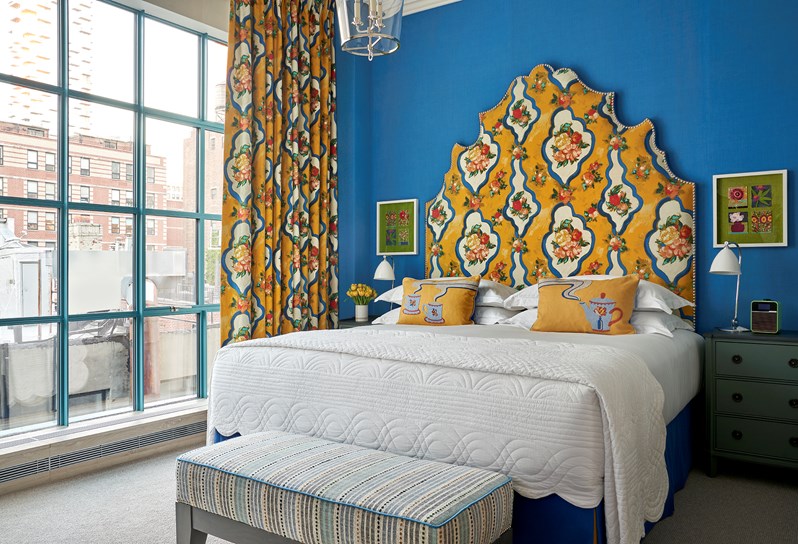 In front of blue wall, there is a bed with a yellow and blue headboard featuring a tropical fruit print which matches the curtains on the left side. There are yellow cushions with a tea cup and teapot detailing on the bed and a blue and white striped ottoman at the end.