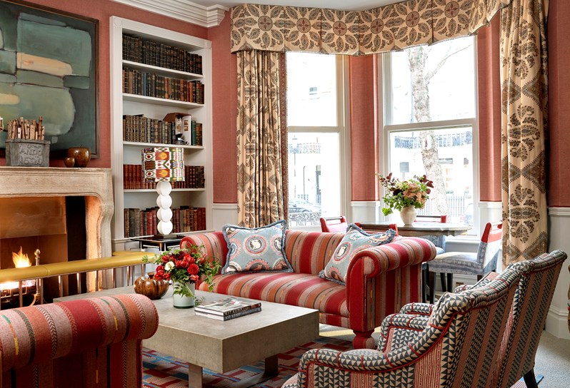 Ample seating in two sofas and two armchairs with a coffee table between, arranged around a fireplace next to a floor to ceiling book case and lots of brightness from big windows.
