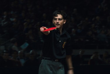 A still from 'Marty Supreme'. A dark haired man with a moustache and glasses points off screen holding a ping pong bat