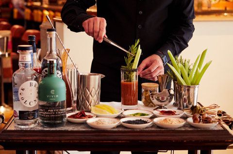 A Bloody Mary trolley displaying various alcohols and garnishes at Refuel Restaurant. A man standing behind places a garnish into a Bloody Mary
