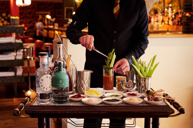 A Bloody Mary trolley displaying various alcohols and garnishes at Refuel Restaurant. A man standing behind places a garnish into a Bloody Mary