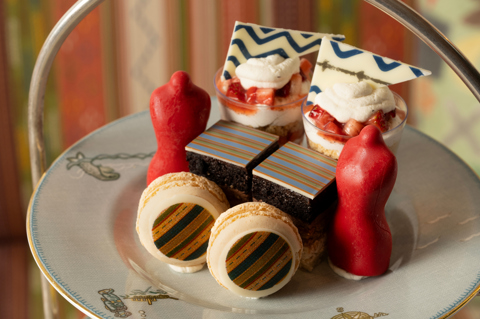 A detail shot of the Kit Kemp Design Thread Afternoon Tea set up at The Whitby Bar. Focusing on the cake stand, there are cakes and macarons decorated with Kit Kemp's famous fabrics, edible mannequins. Sandwiches and scones can be seen out of focus below