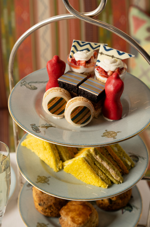A detail shot of the Kit Kemp Design Thread Afternoon Tea set up at The Whitby Bar. Focusing on the cake stand, there are cakes and macarons decorated with Kit Kemp's famous fabrics, edible mannequins. Sandwiches and scones can be seen out of focus below