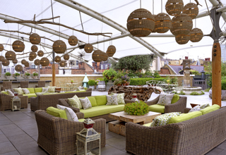 Diagonal shot from the bar of the Roof Terrace at Ham Yard Hotel, it shows the lime green cushioned and wicker sofa seating area with a coffee table in the centre. 