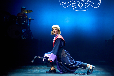 A production shot from Inter Alia. A woman dressed in a judge's wig and robe lunges across the stage holding a microphone stand and looking out in to the audience