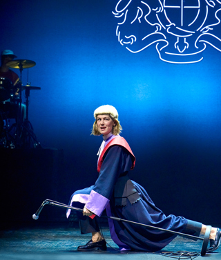 A production shot from Inter Alia. A woman dressed in a judge's wig and robe lunges across the stage holding a microphone stand and looking out in to the audience