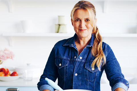 Chef Donna Hay holding baking equipment in a kitchen. She has a large bowl and is surrounded by ingredients
