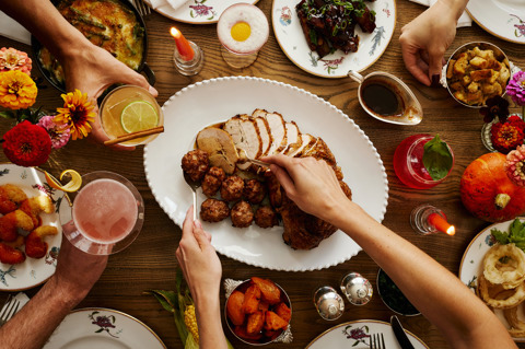 This image is shot from above in Oscar, at Charlotte Street Hotel, to promote the thanksgiving menu friendsgiving. The image shows multiple dishes from the menu including turkey, onion rings, ribs, roast vegetables, mac and cheese and lots of the themed cocktails., with hands passing the items around the table. 