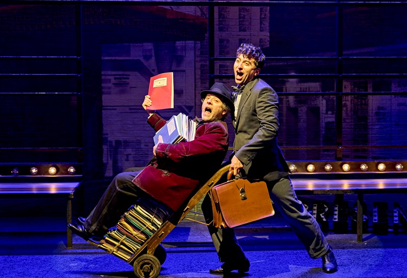 A cast image from the theatre production 'The Producers' to promote the 'The Producers Theatre Package' at Covent Garden Hotel's Brasserie Max. The scene shows one man pushing another man on a trolly, distributing books and singing.
