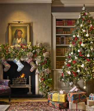 This image shows a beautifully decorated Christmas setting in the library guest area at Haymarket Hotel, featuring a Christmas tree adorned with twinkling lights, red berries, pinecones and ornaments, alongside a roaring fireplace dressed with festive garlands and stockings, with colourful wrapped presents beneath the tree.