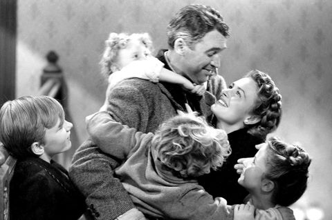 A black and white still image from 'It's a Wonderful Life'. A man stands holding his wife and smiling. His arms extend to hold two young boys. There is a young girl climbing on this back and a young boy to the left of him