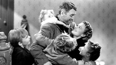 A black and white still image from 'It's a Wonderful Life'. A man stands holding his wife and smiling. His arms extend to hold two young boys. There is a young girl climbing on this back and a young boy to the left of him