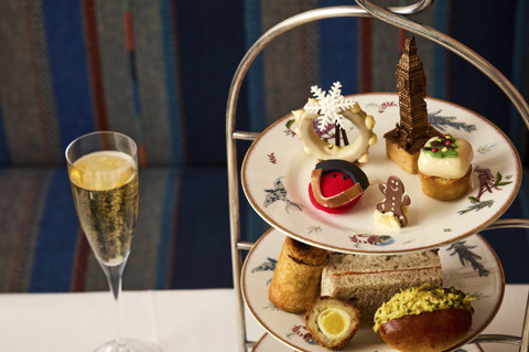 This is close up image of the Christmas London Calling festive afternoon tea offering at Brumus at Haymarket Hotel, the image shows a close up of the plate of cakes offered all in a festive and British theme. There is a glass of champagne next to the stand of afternoon tea.