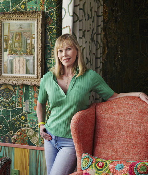 Firmdale Hotels Creative Director Kit Kemp standing her showroom at Kips Bay in New York. She stands in front of a patterned wall leaning on a red armchair