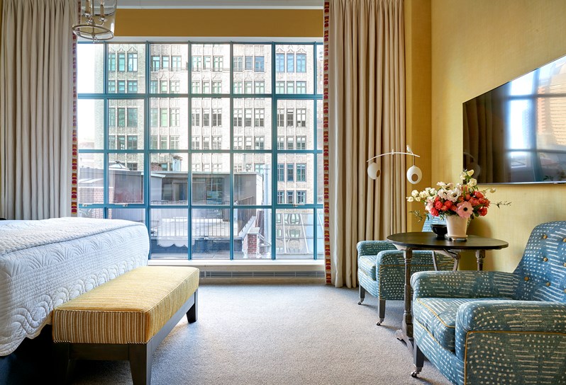 A yellow room, with floor to ceiling windows along the back. On the right, the end of a bed and a yellow ottoman are visible. On the left, two blue and white patterned chairs and a coffee table with a TV hanging above.