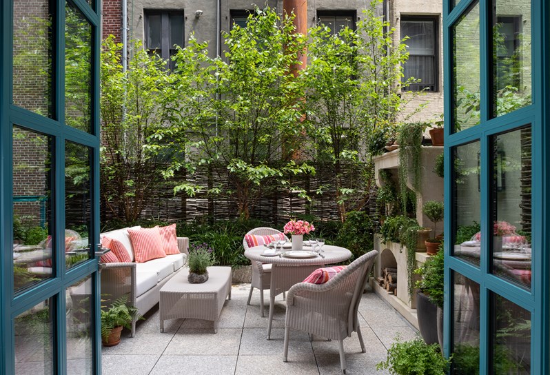 Two blue windowed doors open onto a terrace in the city with a white wicker table and chairs decorated with coral cushions. It is surrounded by several plant pots filled with greenery and at the back of the terrace there is a stone fireplace decorated with plants.