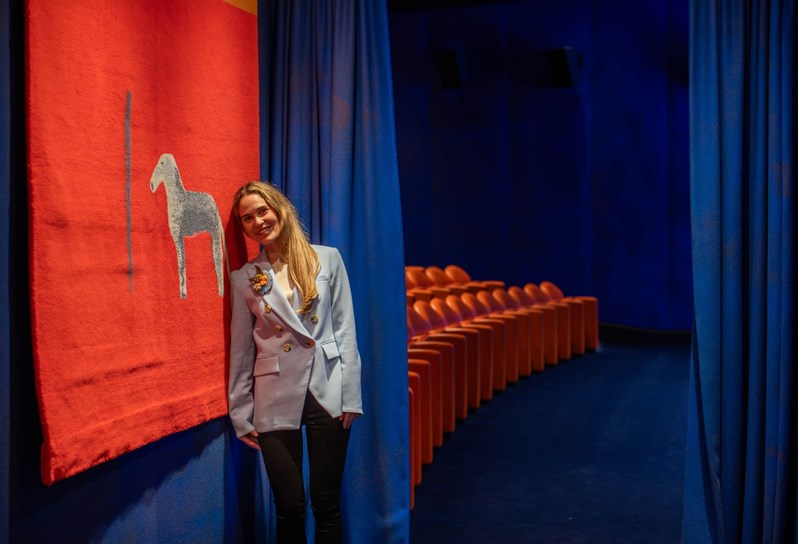 Firmdale Hotels' Art Ambassador and Design Director Willow Kemp stands in a blue blazer and black trousers in The Whitby Hotel's screening room. She is leaning against a blue fabric lined wall with a red printed piece of artwork hanging off it. Behind her, blue curtains are pulled back to reveal rows of orange cinema armchairs to the left