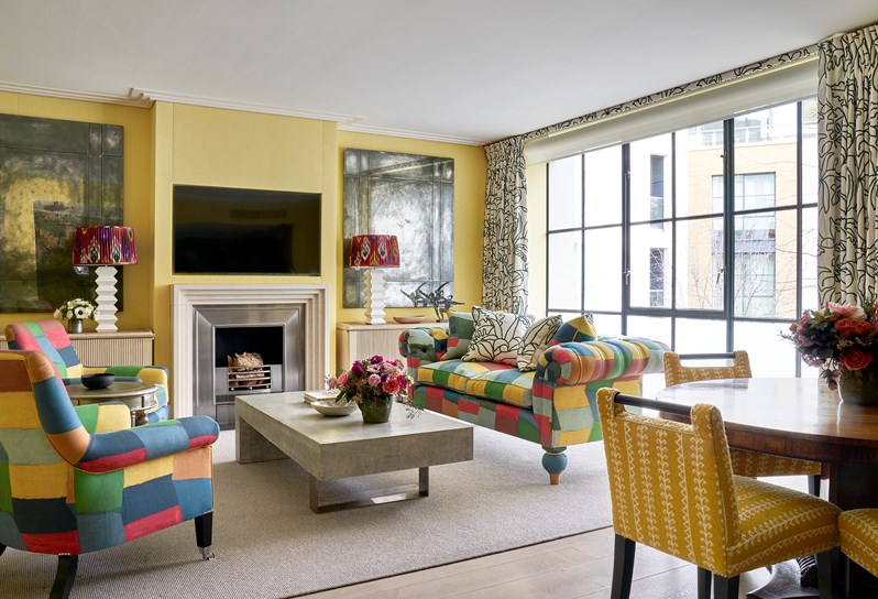 A living room space with a multicoloured sofa and armchairs and a rectangular coffee table. Two mirrors hang either side of the fireplace and a round wooden table sits in the foreground with yellow chairs