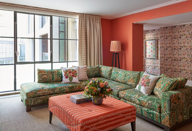 A living room space of the Deluxe Two Bedroom Suite Room 5 at Ham Yard Hotel.  A vibrant living room with red-orange walls, patterned green furniture, and eclectic décor floral arrangements, and framed textile art above the fireplace.