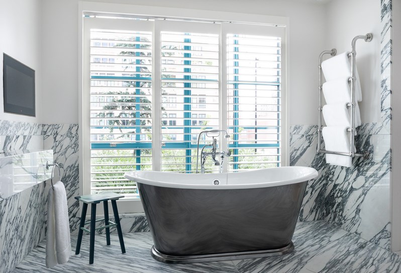 Wide shot of a marble bathroom. A large freestanding bath sits in the centre in front of large window. To the left, there is a TV on the wall and to the right, there is a towel rail.