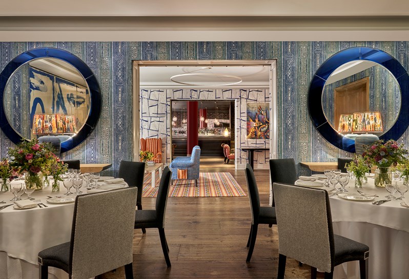 An image of multiple round dining table set up in the Indigo Room, there are white table cloths, floral bouquets in the centre and candles. each place around the table has a placemat, plate, cutlery, glassware and napkins ready for dining. private events room at The Soho Hotel.