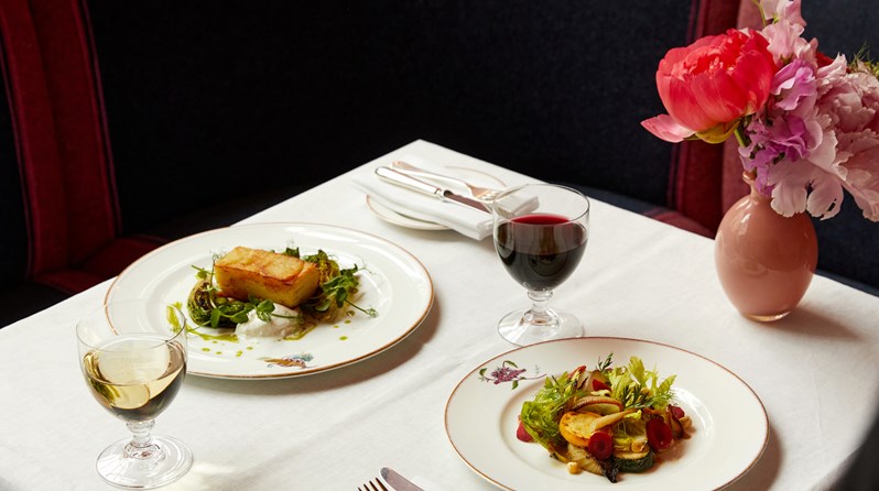 A dining table set up for two in front of booth seating. A plate with a layered potato and salad on and a plate with vegetable salad, a glass of red wine and white wine.