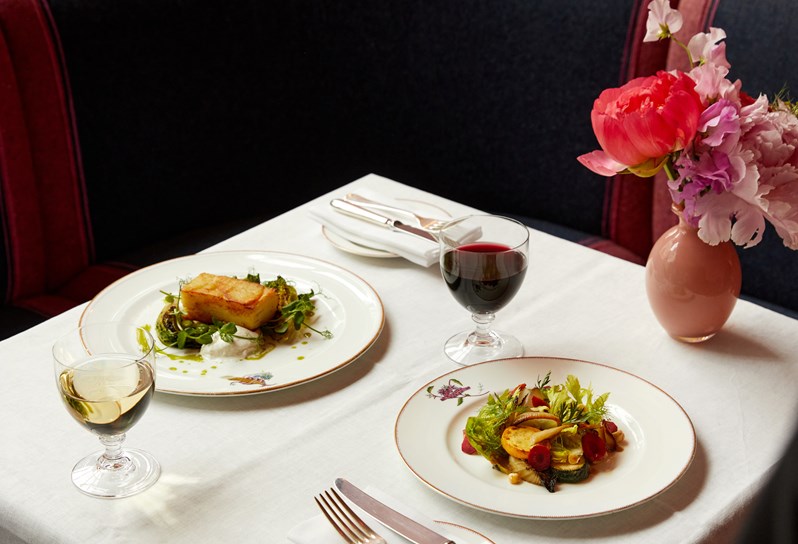 A dining table set up for two in front of booth seating. A plate with a layered potato and salad on and a plate with vegetable salad, a glass of red wine and white wine.
