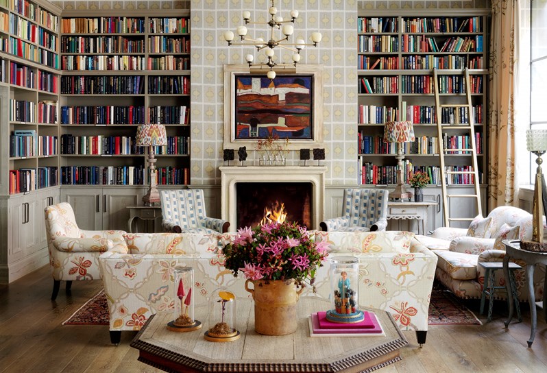 A view of Ham Yard Hotel's Library from the entrance, showing a table with artwork and fresh flowers in the foreground, backed by a seating area with sofas and armchairs in floral and neutral colors and patterns, arranged in front of a fireplace with book filled walls on both sides.