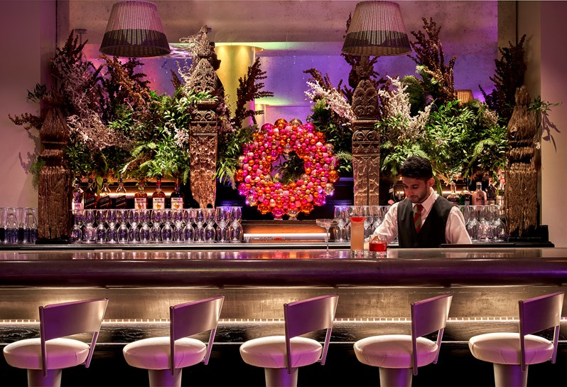 An overview of the bar in the pool area. It is very festively decorated with wineglasses along the table and and a wreath ring made from baubles in the centre. There are 5 bar stools alongside the bar and the bartender can be seen making two cocktails.
