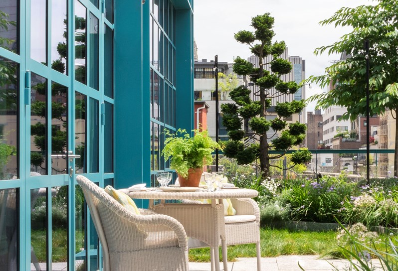 A wraparound landscaped terrace balcony. Plants and grass around the outskirts and a small patio in the middle with white woven outdoor dining chairs and table.