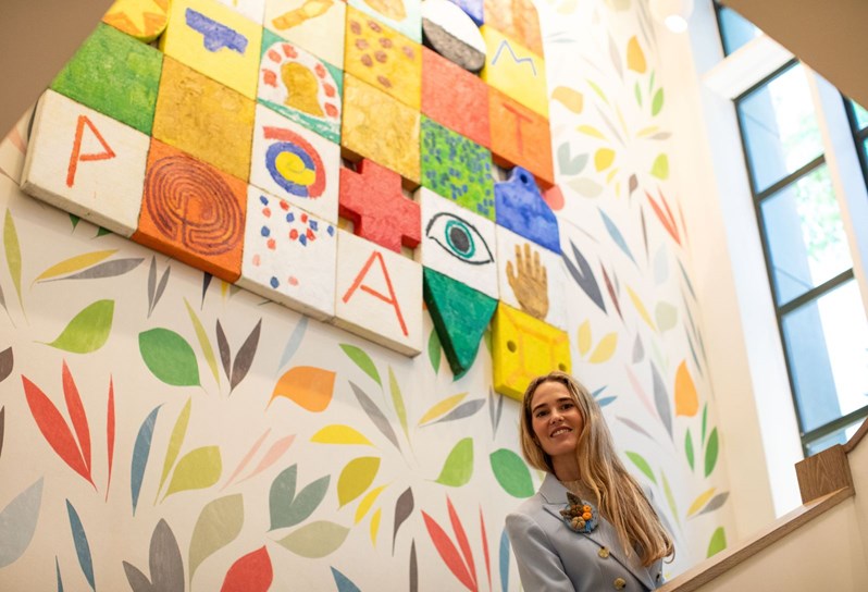 Firmdale Hotels Art Ambassador and Design Director, Willow Kemp is seen peering over a banister on a staircase at The Whitby Hotel looking towards the camera below. Behind her the wall is covered in a colourful leaf pattern and a coloured grid artwork hangs above.