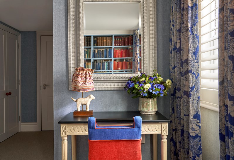 Desk area shot of room 15, the Marylebone Room at Dorset Square Hotel. We see a red and blue chair in front of a white desk with a doggy lamp on top of it. The desk sits beneath a large mirror