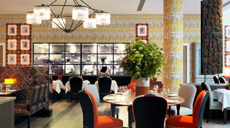 Tables beautifully set for dining in the quirky and playfully decorated restaurant, looking in toward the open plan kitchen with large windows, seeing three chefs at work.