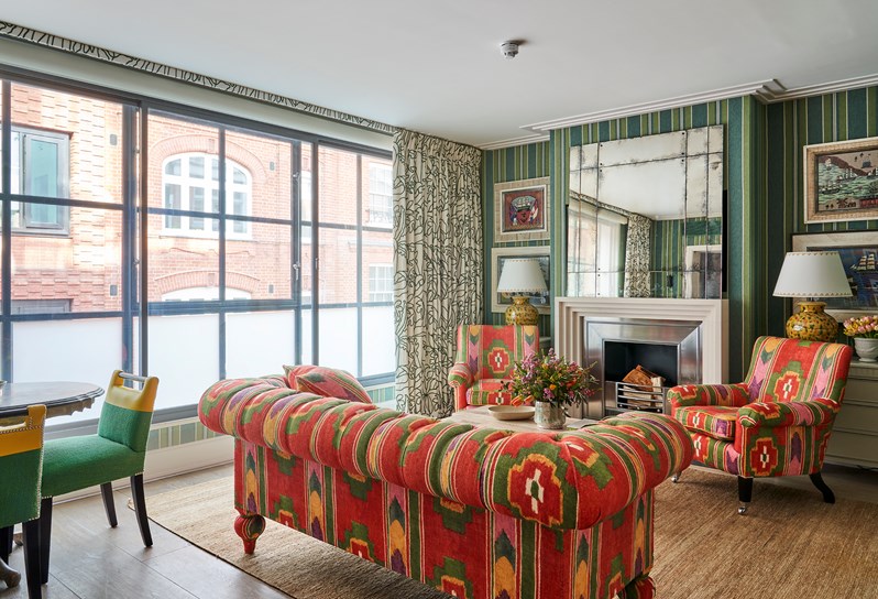 A spacious lounge area in an open plan suite, boasting floor to ceiling windows with a seating arrangement in front; a  comfortable sofa, a wooden coffee table with fresh flowers and a dinning table. Art works hang either side of the fireplace